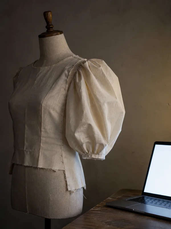 A linen dress form wears an unfinished natural-coloured toile bodice with one large gathered puff sleeve.