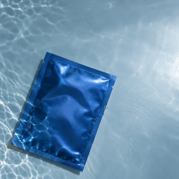 A plain cobalt-blue foil sachet lies diagonally on a pale blue underwater surface, with bright water caustics crossing the scene and a small bubble beside its…