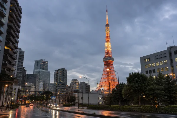 Tokyo Tower glows orange above surrounding city buildings beneath a blue-grey evening sky, with a wet roadway and vehicle lights in the foreground.
