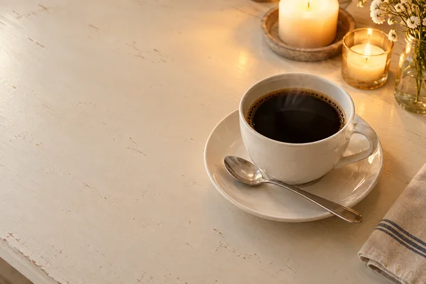 A white ceramic cup of black coffee sits on a matching saucer on a pale cream tabletop, with a stainless-steel teaspoon across the saucer.