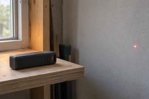 A compact charcoal-grey laser measure rests on a layered plywood ledge in an unfinished workshop, aimed toward a pale plaster wall where a small red dot is…