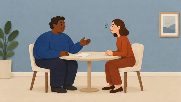 A dark-skinned coworker in a blue sweater gestures across a cream table toward a peach-skinned coworker in rust clothing, who leans forward with hands clasped.