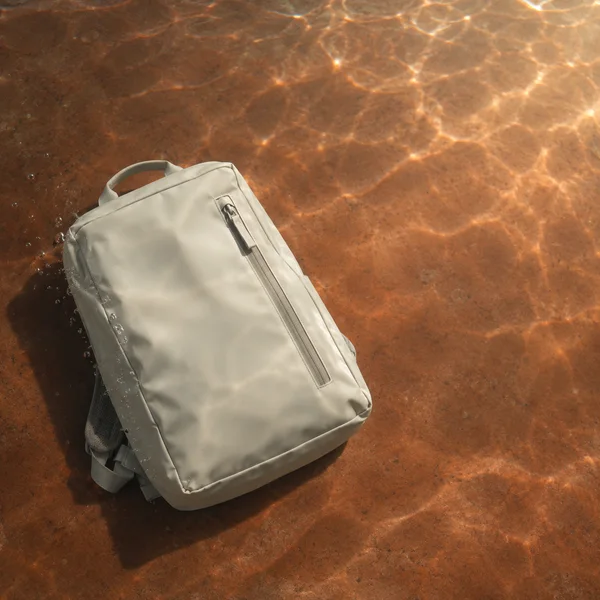 An ivory rectangular backpack lies diagonally on a terracotta-colored floor beneath clear water, with rippling light patterns and small bubbles along one side.