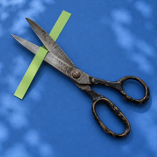 An aged pair of steel scissors with chipped black handles lies open diagonally on a blue surface, crossing a narrow lime-green paper strip; soft mottled…