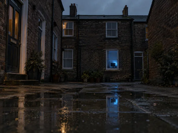 A wet brick residential courtyard at dusk, with a blue-lit ground-floor window and its reflection in a large foreground puddle.