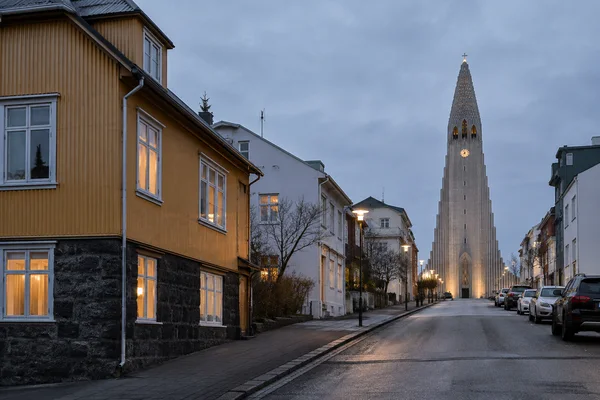 A quiet Reykjavik residential street at dusk, looking uphill past an ochre corrugated-metal townhouse with glowing windows toward Hallgrimskirkja beneath an…