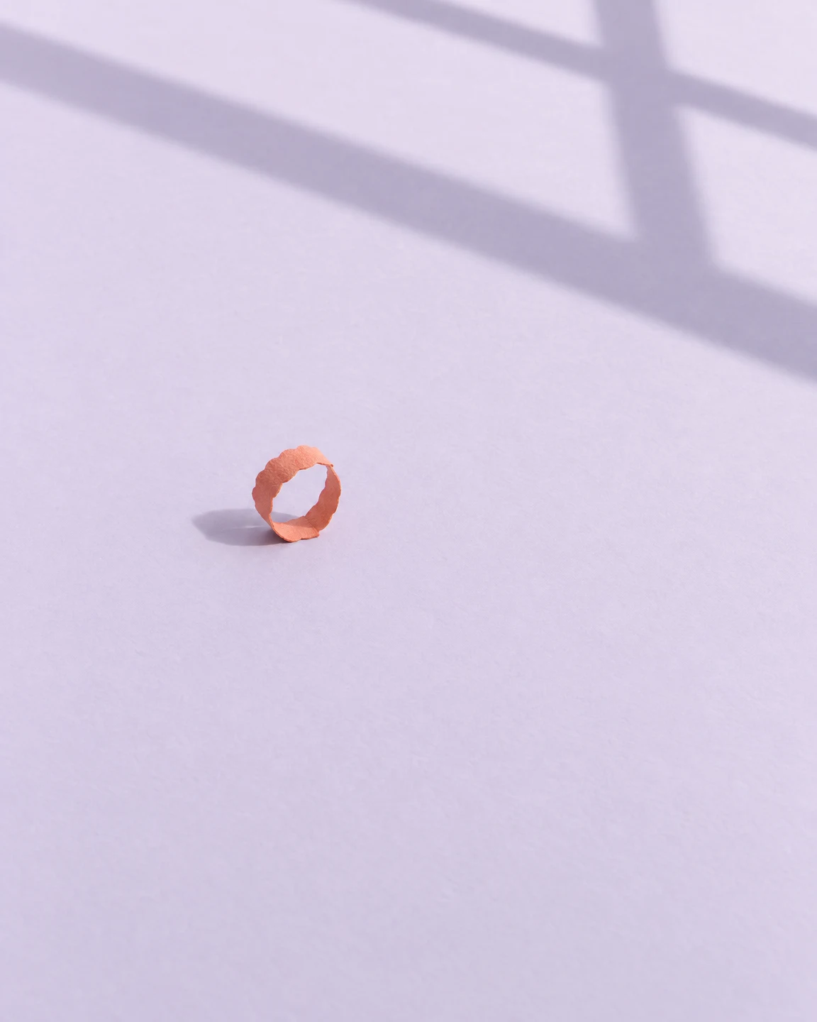 A small coral-orange scalloped paper loop stands on a pale lilac surface beneath a broad diagonal window-frame shadow.
