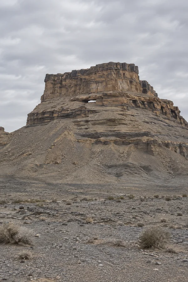 A layered, flat-topped sandstone mesa with a shallow notch beneath one ledge rises above a gravelly desert plain dotted with sparse shrubs under a cloudy grey…