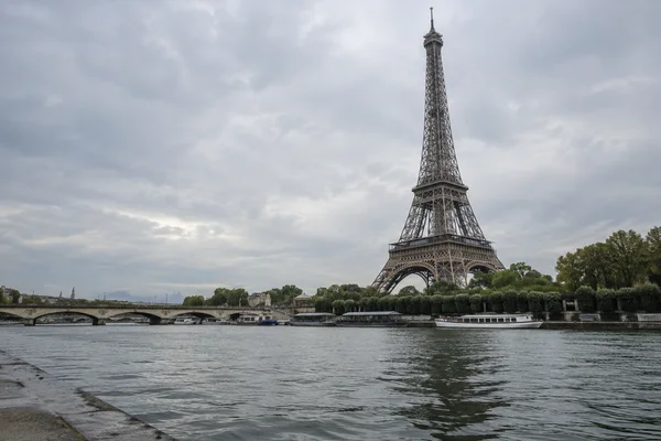 The Eiffel Tower rises above the tree-lined Seine embankment beneath a broad overcast sky, with boats along the opposite bank, a bridge to the left, and…
