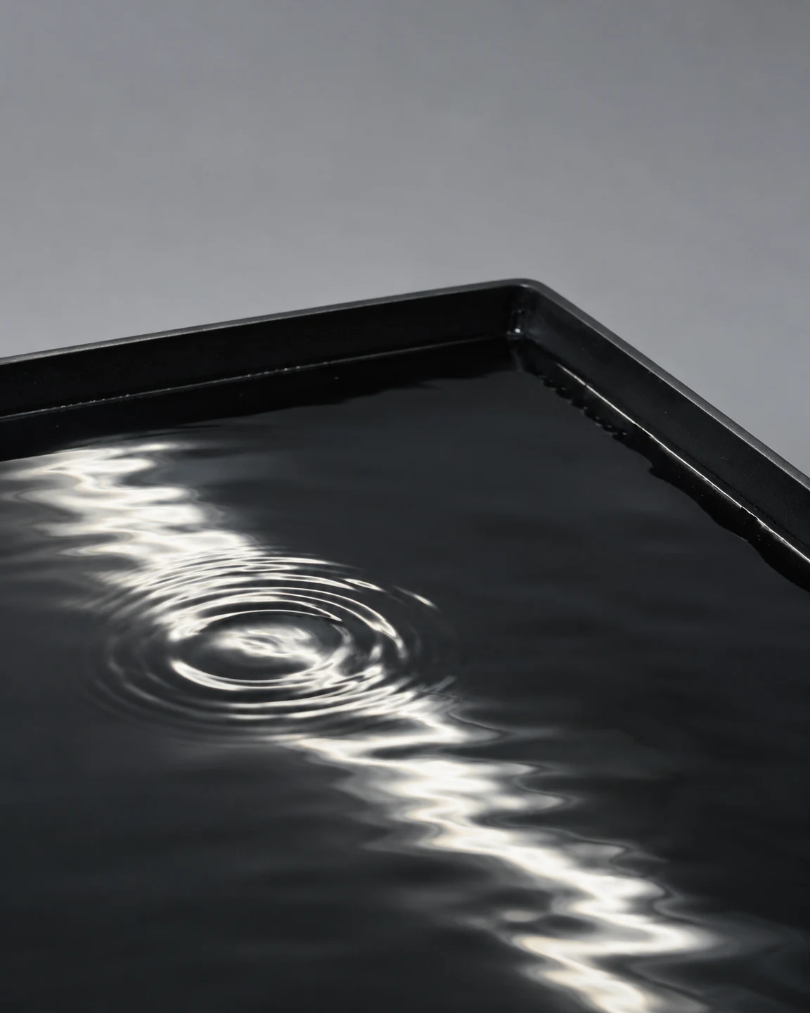 A shallow black rectangular tray of dark water sits against a cool grey background, with a bright winding reflection crossing the surface and circular ripples…