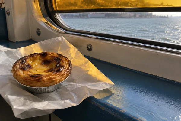 A blistered custard tart in a crimped silver foil case sits on folded white paper on a blue-painted ferry ledge, with river water visible through the window…