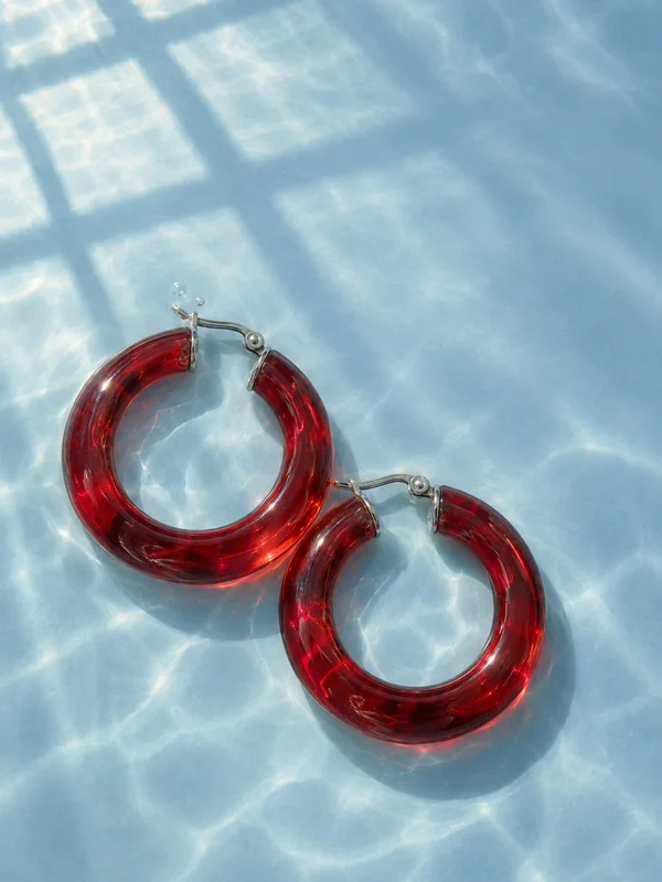 Two chunky translucent scarlet hoops with silver closures and two small bubbles rest on a pale-blue underwater surface, beneath broad window-shaped light and…