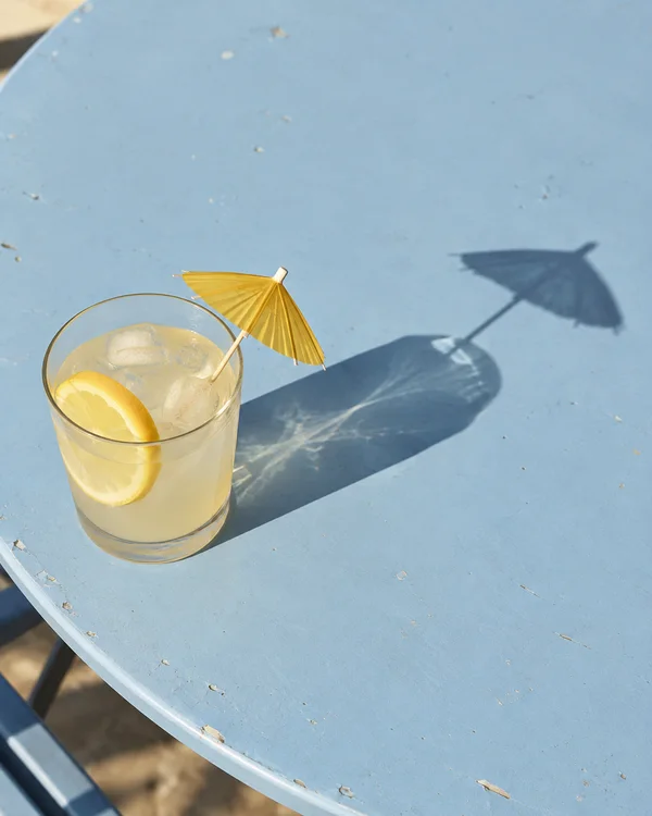 A clear glass of pale lemonade with ice and a lemon slice sits on a worn pale-blue café table.