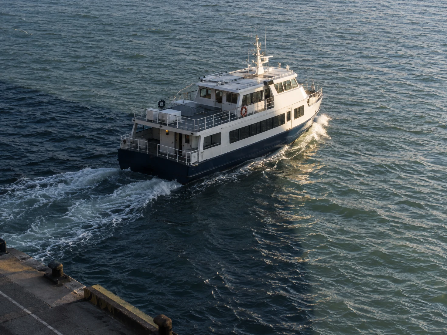 An elevated view of a white two-deck ferry with a navy lower hull moving diagonally across choppy harbor water.