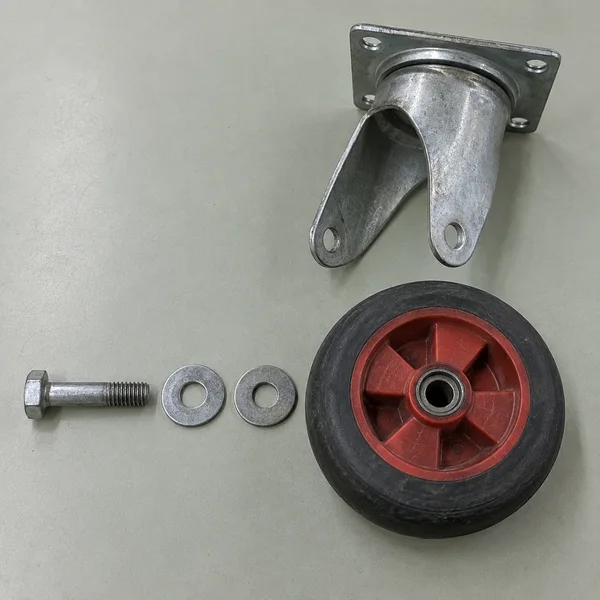Overhead view of a dismantled utility-trolley caster on a pale grey-green desk: a red wheel with a black tire sits below its galvanized-steel fork, while a…