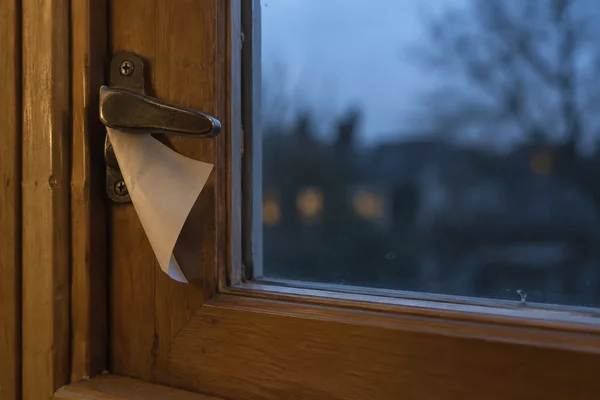 A blank cream sheet is caught beneath a dark metal latch on a honey-coloured wooden window frame, with cool blue-grey glass filling the right side of the image.