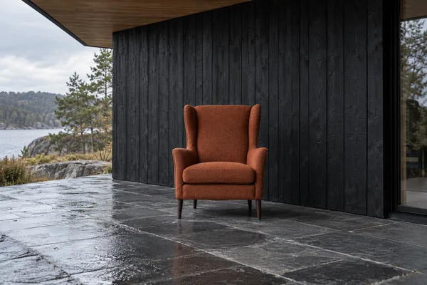 A rust-colored wingback armchair stands on dark slate paving beside a black vertical timber wall beneath a timber canopy, with a lake, rocky shoreline, and…