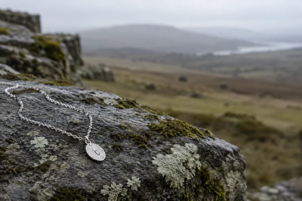 A fine silver chain with a small oval pendant rests on a damp, lichen- and moss-speckled grey stone ledge, with a softly blurred grey-green moorland and pale…