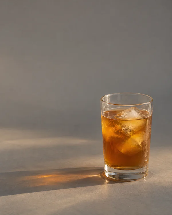 A clear straight-sided glass of amber iced tea with ice sits on a cool-grey surface, lit warmly from the left and casting a long soft shadow; broad empty space…