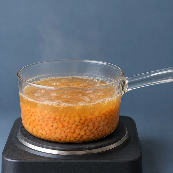 A clear glass saucepan filled with orange-red lentils and golden-orange liquid sits on a dark electric hotplate against a slate-blue backdrop.