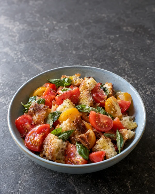 A pale-blue ceramic bowl of panzanella with red and golden tomato wedges, browned bread pieces and basil sits on a charcoal stone tabletop, with open surface…