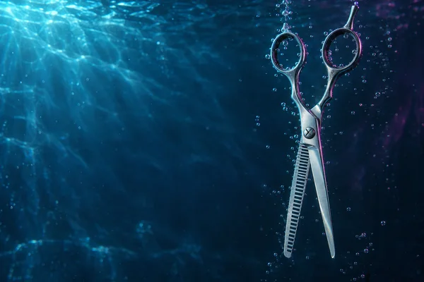 A stainless-steel hair-thinning shear hangs open underwater on the right, with its toothed blade facing a smooth blade; bubbles surround it against a blue…