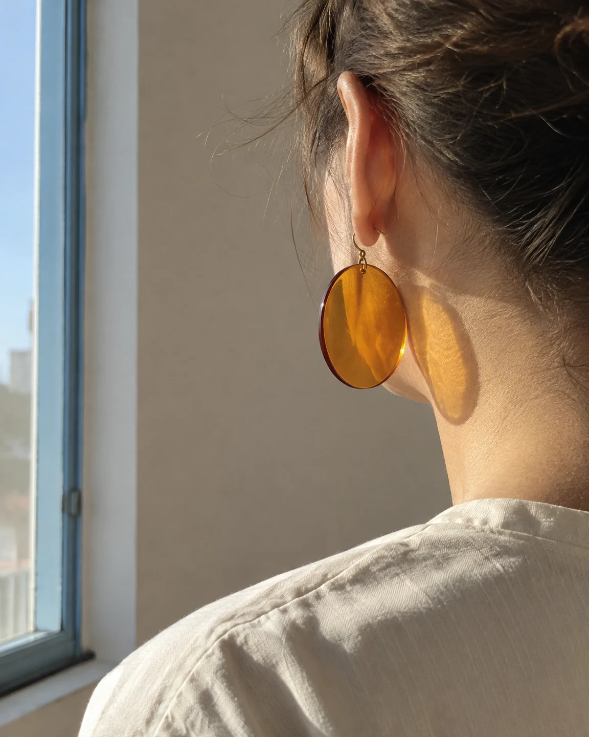Rear three-quarter close-up of a person wearing a large translucent amber disc earring beside a sunlit neck, with a pale blue window frame at left and a plain…