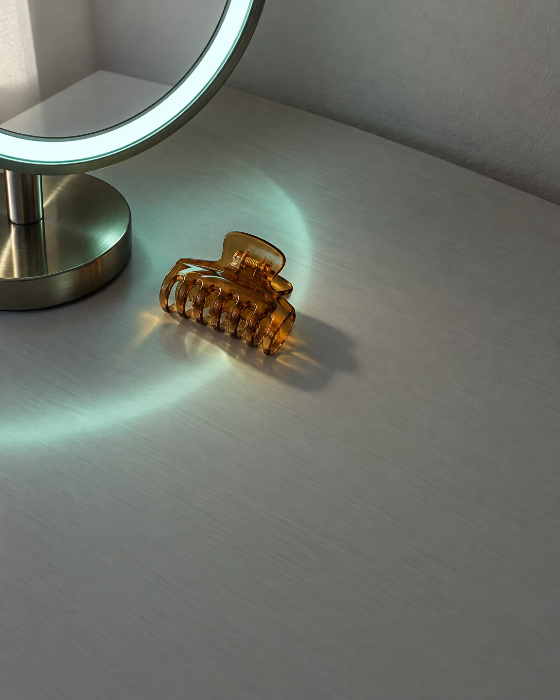 An amber translucent claw hair clip rests on a pale grey, subtly textured tabletop beside a cropped circular mirror with a glowing cyan-green rim.