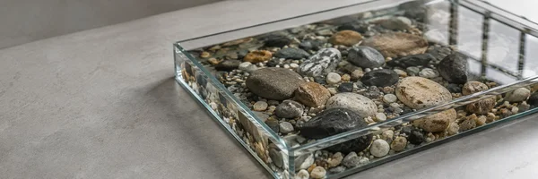 A shallow rectangular glass tray filled with rounded river stones sits diagonally on a warm-gray surface, with open space to the left and window reflections…