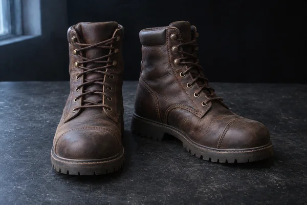 Two dark brown leather lace-up work boots stand on a scuffed charcoal bench, one facing forward and the other angled outward, with a cool-lit window visible at…