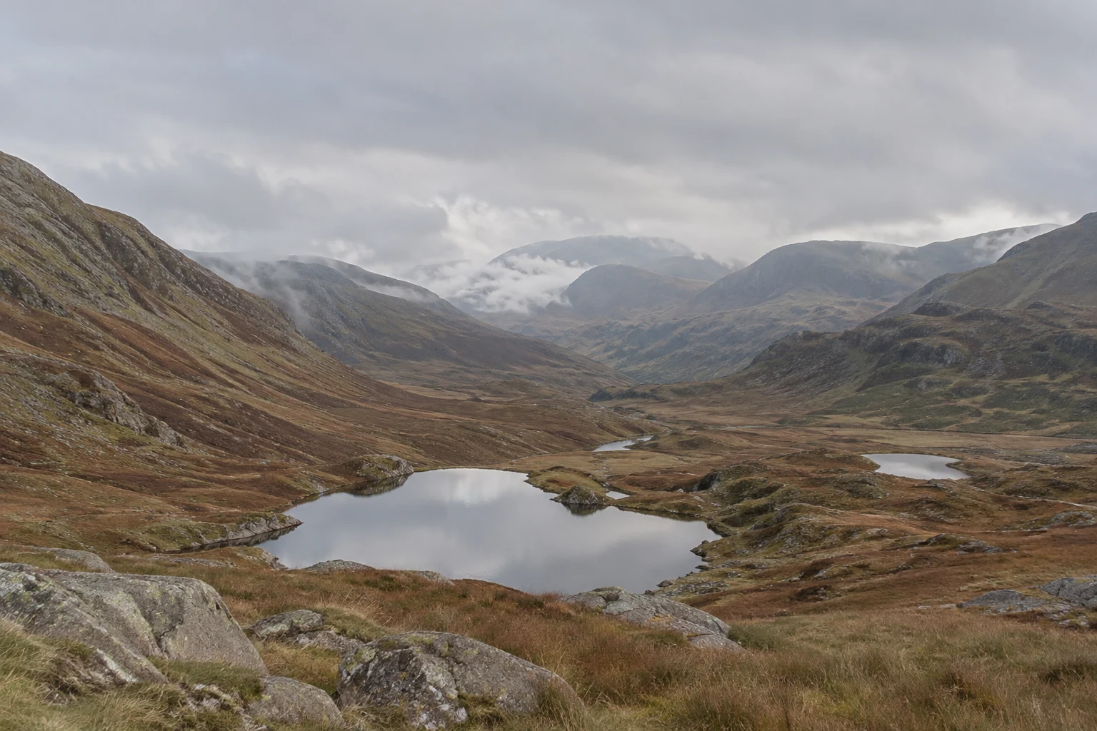 A still mountain tarn reflects the overcast sky in a broad valley, with rust-brown grass, grey foreground boulders, layered ridgelines and low cloud.