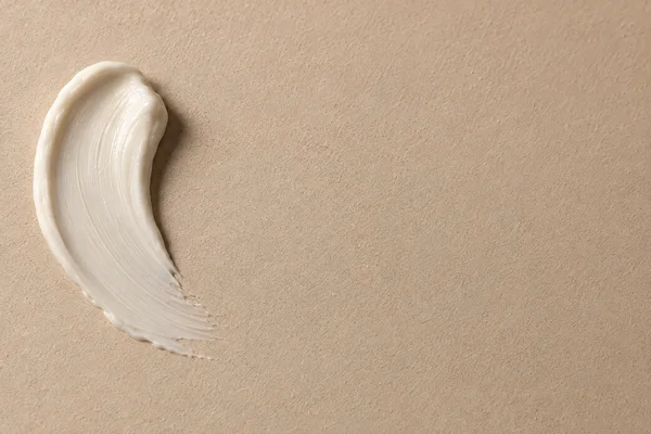 A curved swipe of ivory cream with fine parallel spatula ridges and a rounded raised edge sits on a warm sand-colored textured surface, leaving open space to…
