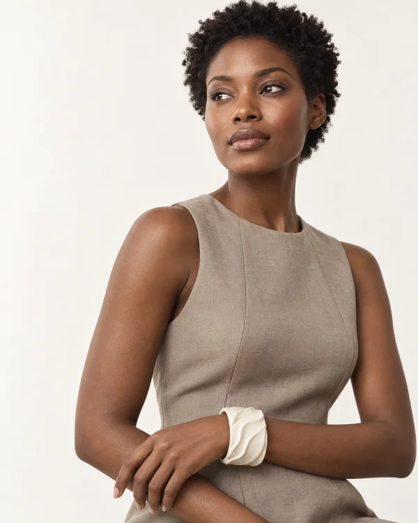 A woman with short natural curls wears a sleeveless taupe top and a broad ivory ridged cuff, resting one forearm across her waist against a warm-white studio…