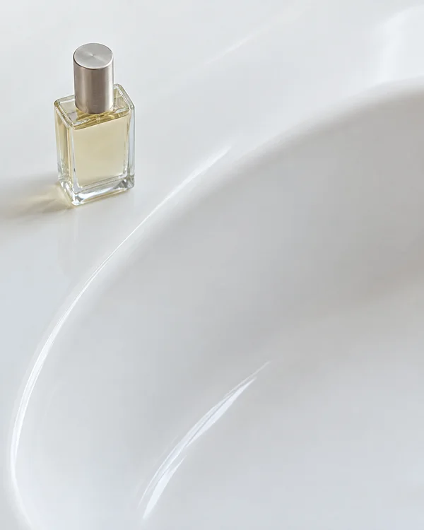 A clear rectangular perfume bottle with pale golden liquid and a brushed-silver cap stands on a white porcelain basin; the basin’s curved rim sweeps diagonally…