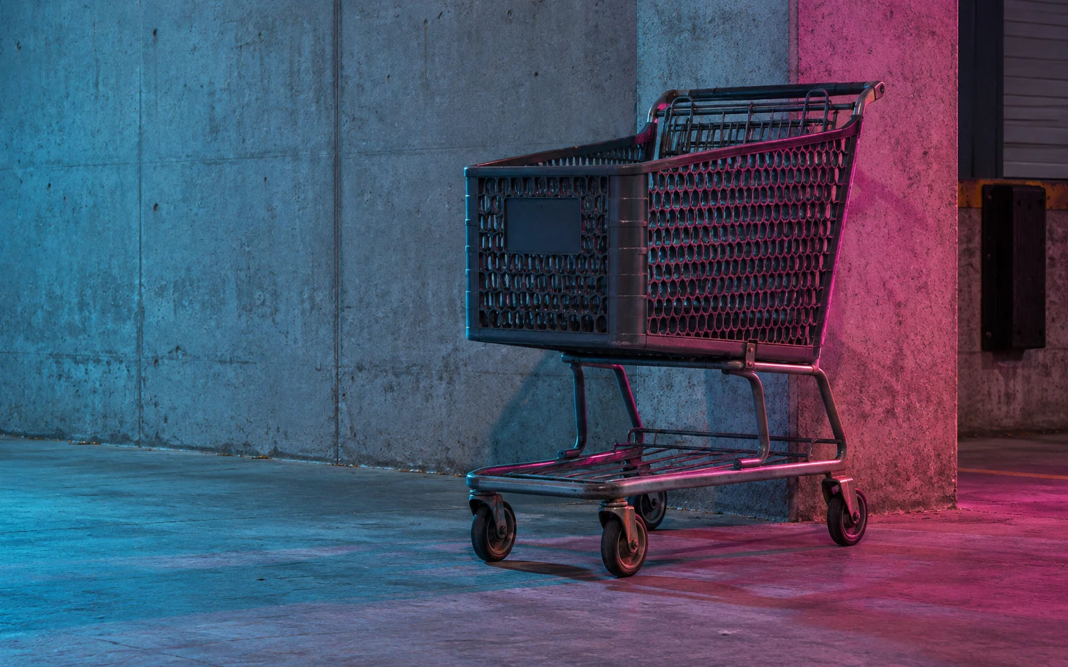 An empty dark shopping cart stands on a concrete floor beside a broad concrete wall and pier, lit with cyan and magenta highlights.