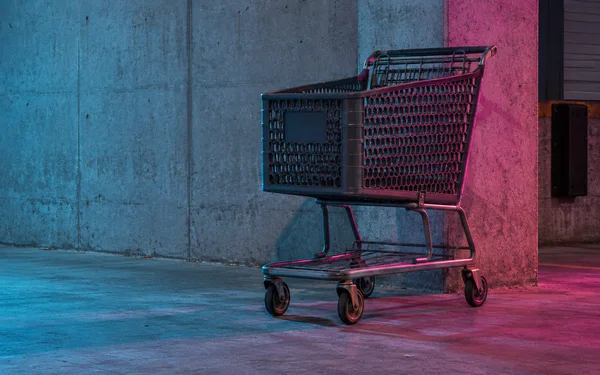 An empty dark shopping cart stands on a concrete floor beside a broad concrete wall and pier, lit with cyan and magenta highlights.