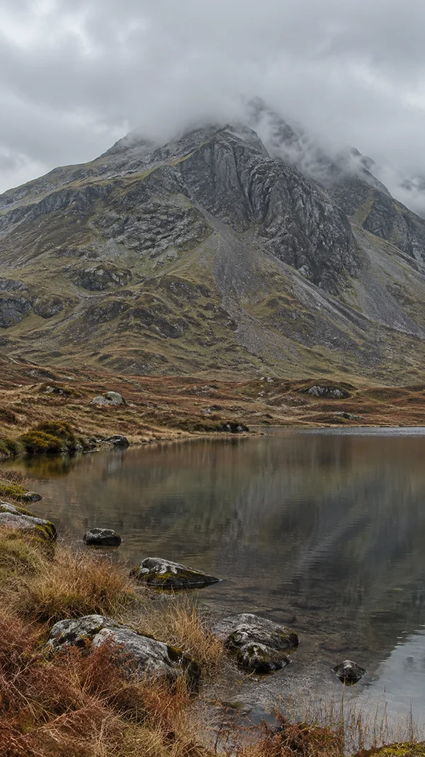 A steep, dark rocky mountain rises into layered grey cloud above a still tarn, with muted brown grasses, bracken and stones along the near shore.