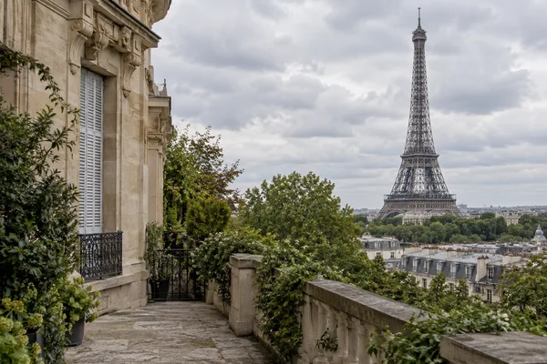 A stone terrace beside a pale limestone townhouse overlooks Paris rooftops and the Eiffel Tower beneath an overcast sky.
