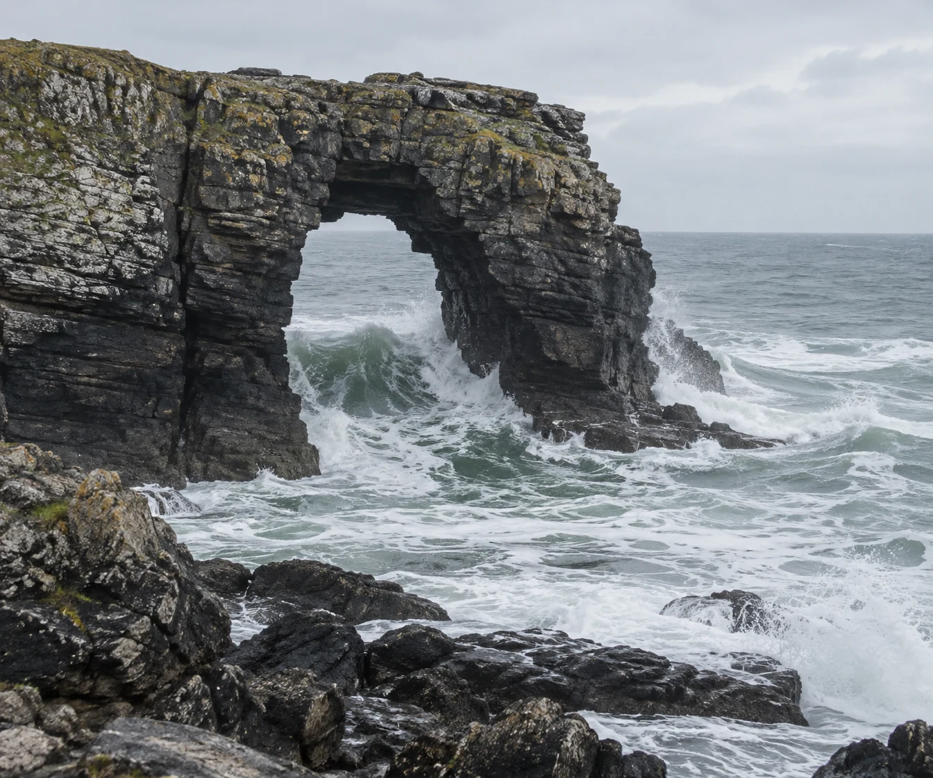 A dark, layered sea arch rises from the rocky coast as a grey-green swell breaks through it, churning into white foam around the rock base.