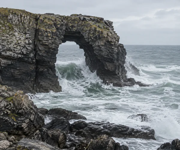 A dark, layered sea arch rises from the rocky coast as a grey-green swell breaks through it, churning into white foam around the rock base.