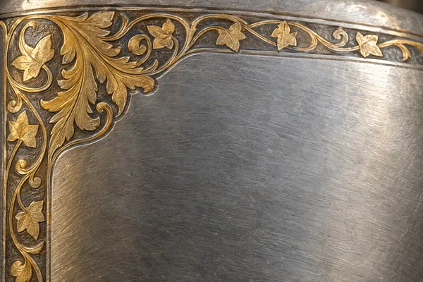 An extreme close-up of a brushed silver surface with gold-toned engraved foliage framing the upper-left corner and continuing along the top and left edges.