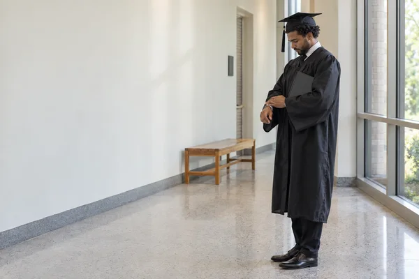 A graduate in a black mortarboard and gown looks down while adjusting a cuff in a bright college corridor, with a closed dark folder tucked under one arm and a…