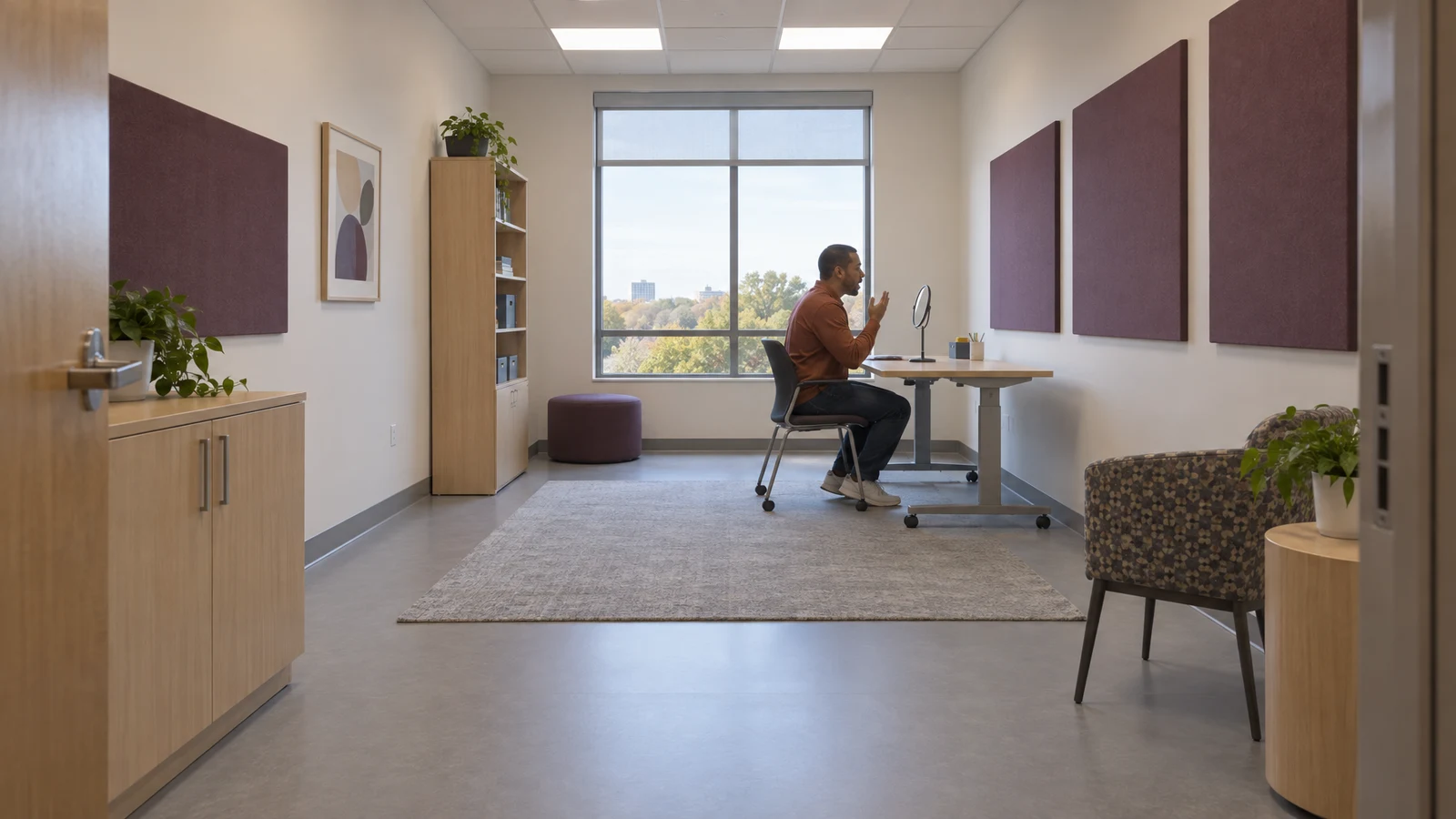 A seated adult in a rust-colored shirt practices speaking toward a small tabletop mirror in a spacious therapy room, with plum acoustic panels, a large window…
