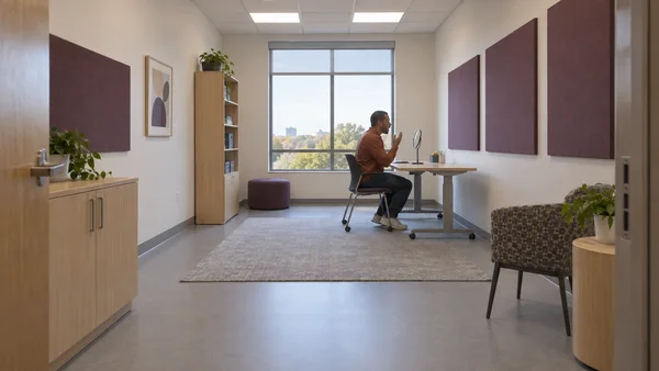 A seated adult in a rust-colored shirt practices speaking toward a small tabletop mirror in a spacious therapy room, with plum acoustic panels, a large window…