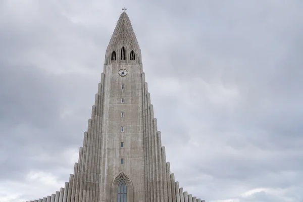 A frontal view of Hallgrímskirkja’s tall concrete tower, with its clock, pointed crown and cross above stepped wings against a cloudy grey sky.
