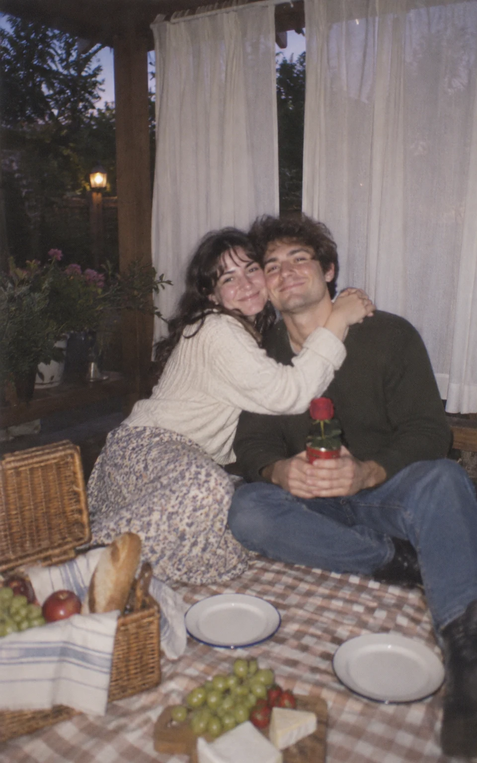 A young woman hugs a young man seated on a checked blanket beneath white curtains at dusk.