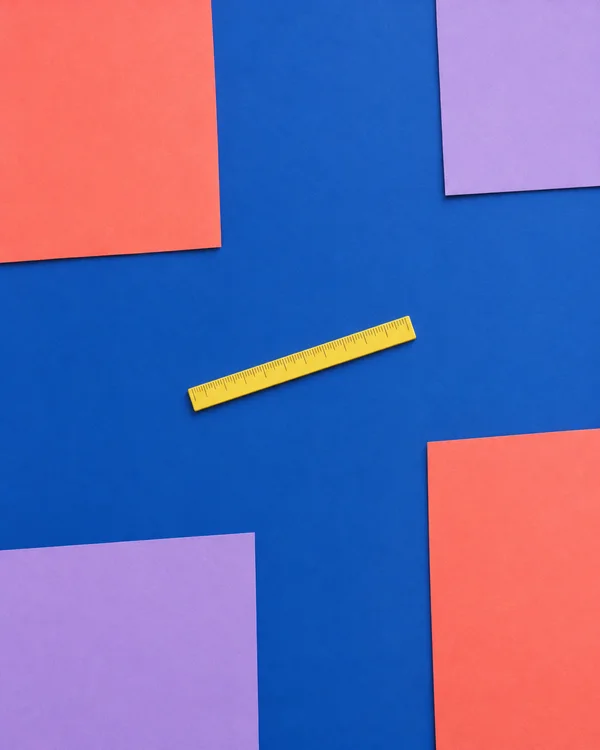 A slim yellow ruler with dark measurement ticks lies diagonally across a matte cobalt-blue surface, framed by cropped coral and lilac paper panels at the edges.