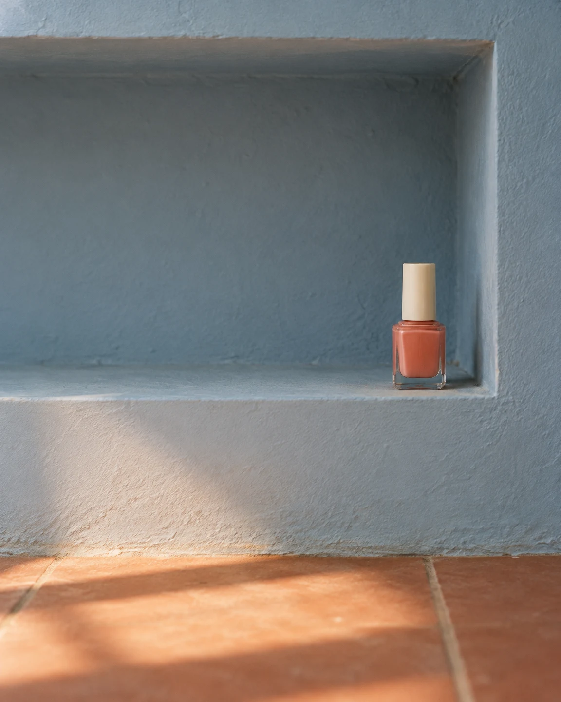 A small clear bottle of coral nail polish with an ivory cap stands at the right end of a pale blue recessed shelf, above a terracotta tile floor lit by warm…