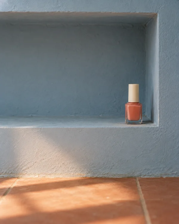 A small clear bottle of coral nail polish with an ivory cap stands at the right end of a pale blue recessed shelf, above a terracotta tile floor lit by warm…