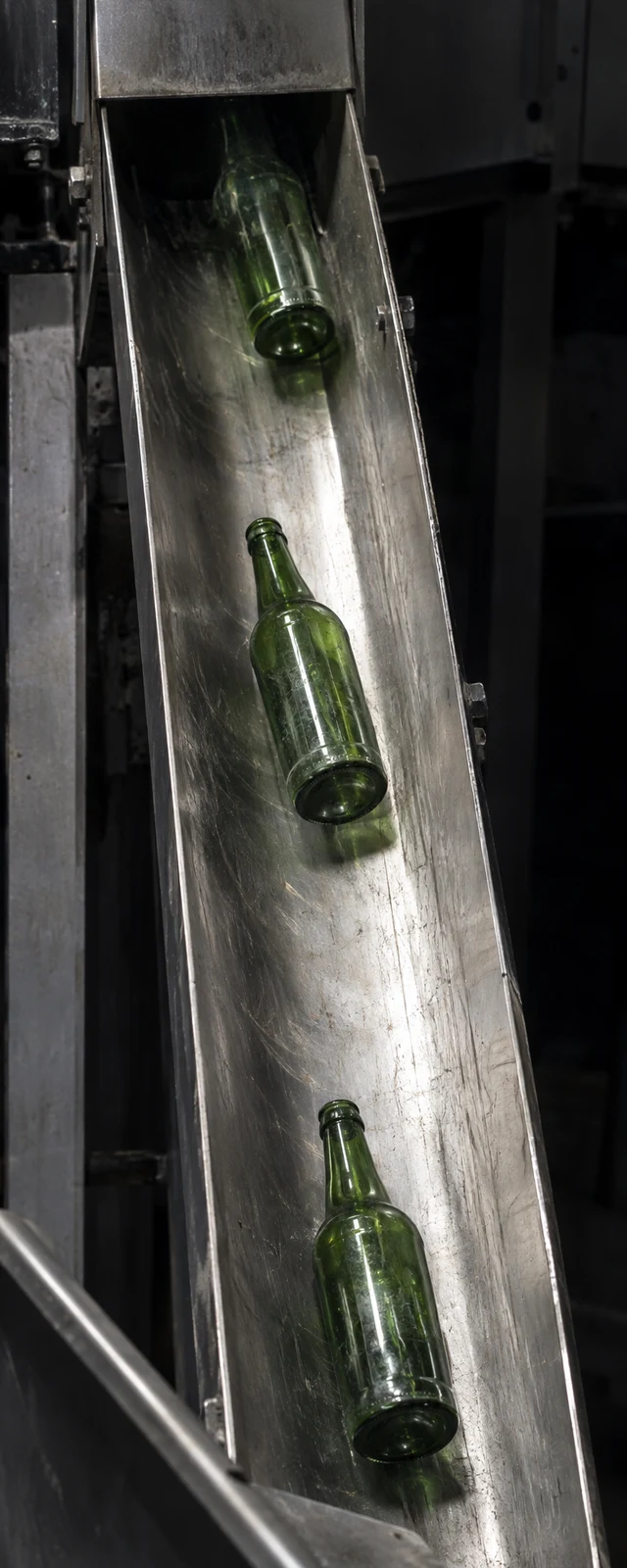 Three green glass bottles appear at separated positions on a scratched stainless-steel chute in a dark industrial bay.